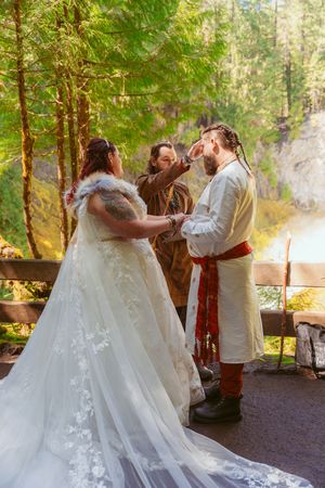 A medieval-themed wedding ceremony takes place on a forested path with sunlight streaming through trees.