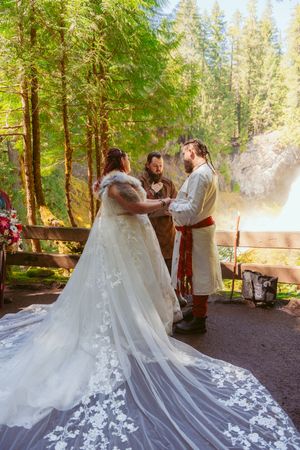 A bride in a flowing white gown exchanges vows during an outdoor medieval-themed ceremony.