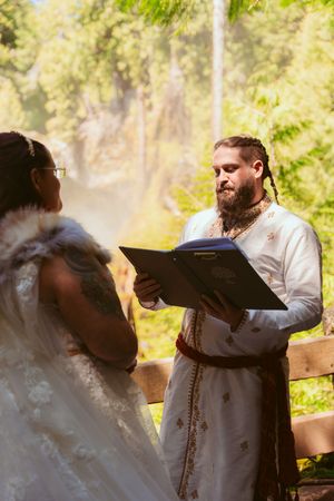 An outdoor wedding ceremony with officiator reading from book near waterfall.