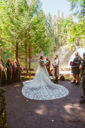 Outdoor forest wedding ceremony with attendees in medieval attire and a dramatic lace train catching sunlight.