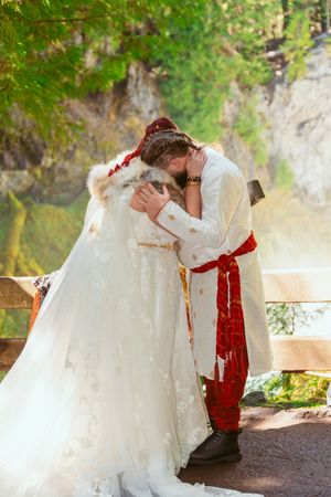 A loving wedding couple shares intimate kisses in the sunlit woods wearing white dress and red accents.