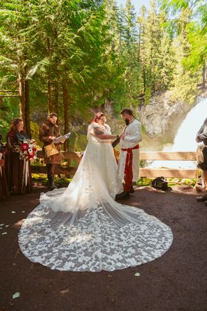A medieval-themed wedding ceremony takes place by a waterfall with a bride wearing an intricate lace train dress.