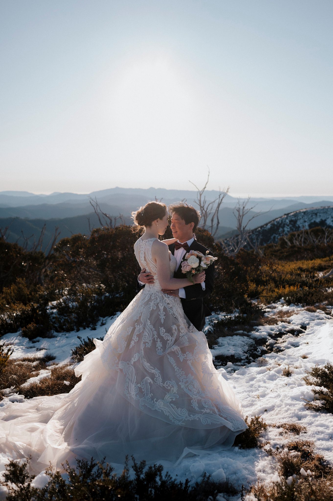 Snowy mountain wedding photo with a flowing lace wedding dress against a dramatic mountain landscape backdrop.