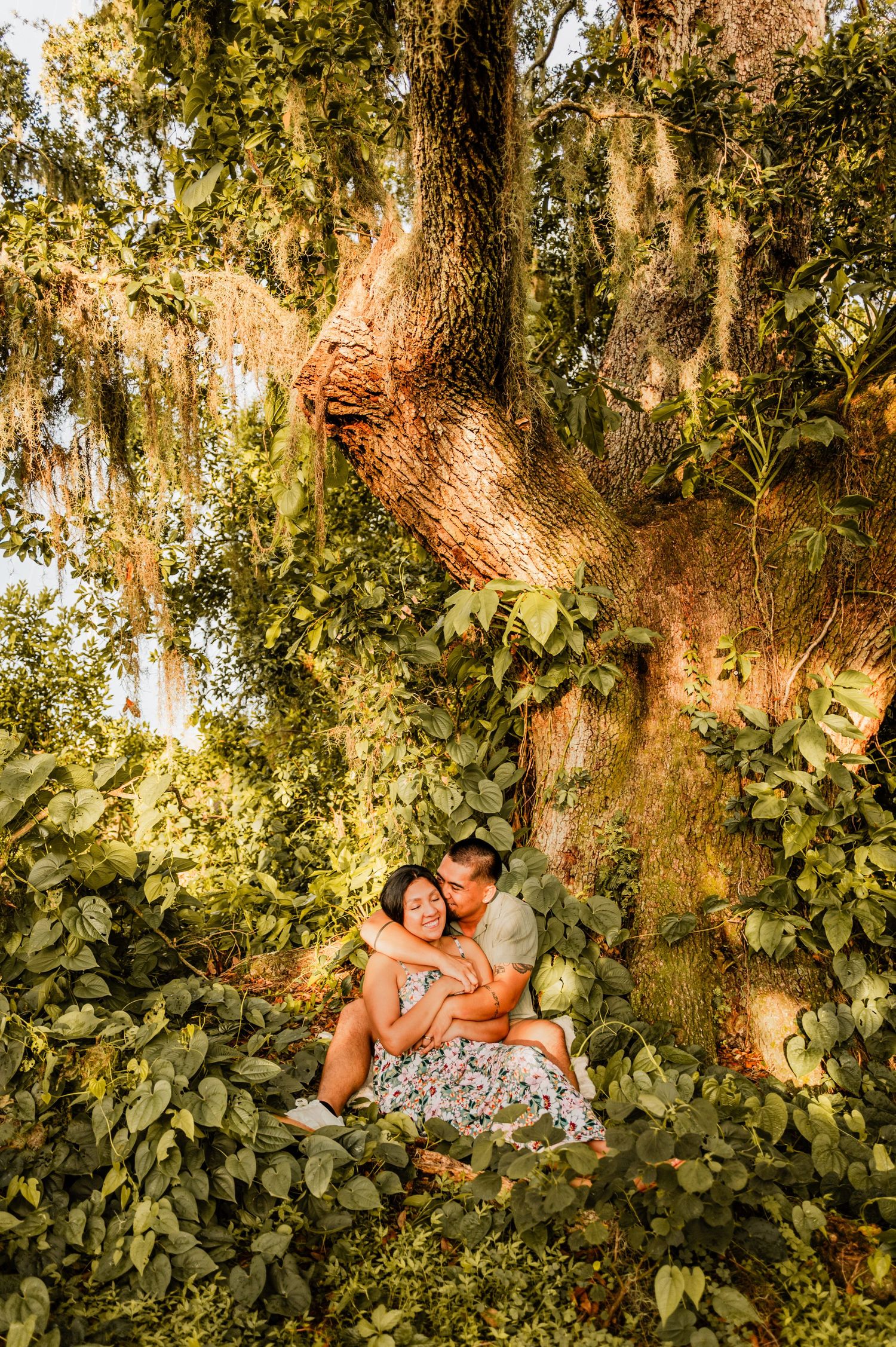 A couple cuddling beneath a large oak tree covered in Spanish moss at sunset.