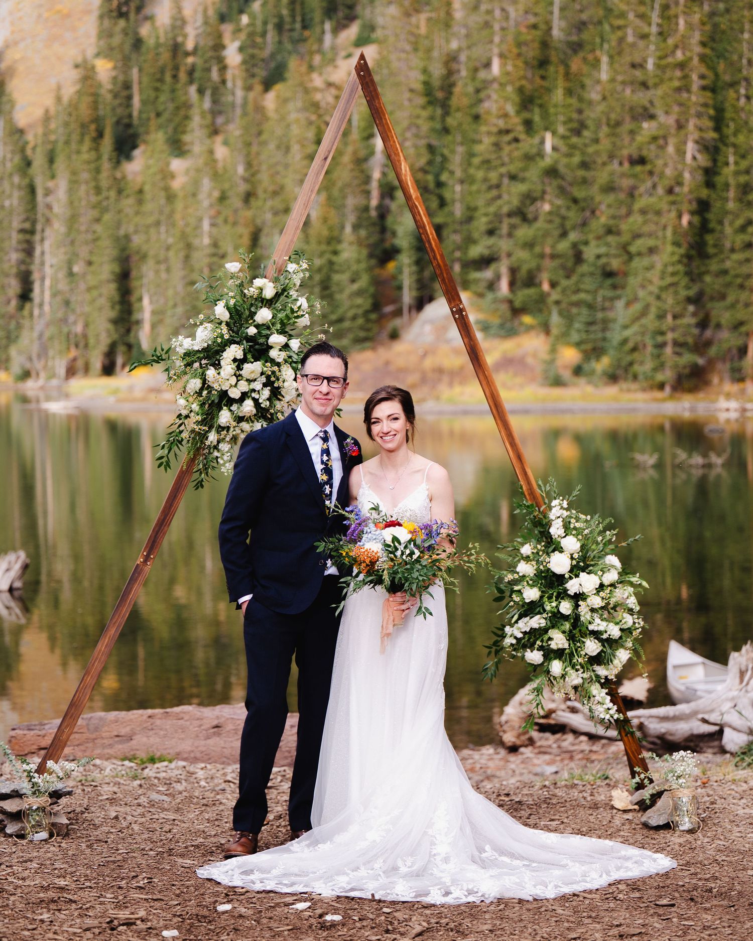 A scenic mountain wedding ceremony takes place beside an alpine lake surrounded by evergreen forests and rugged peaks.