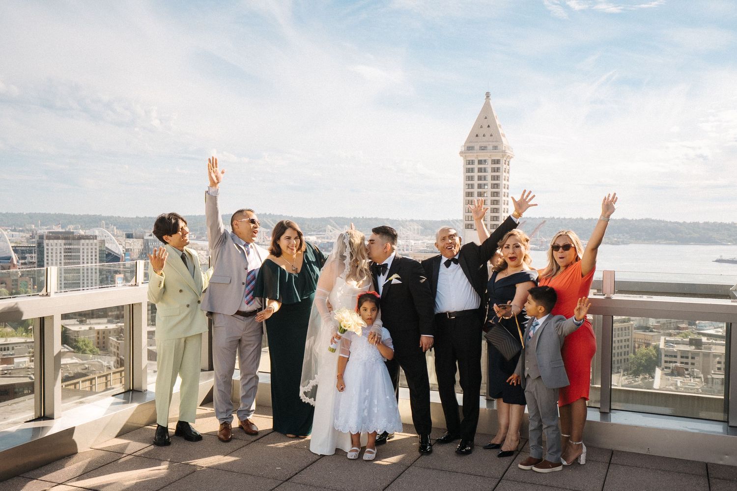 A celebratory group poses joyfully with raised arms on a rooftop terrace overlooking Seattle's Smith Tower and waterfront.
