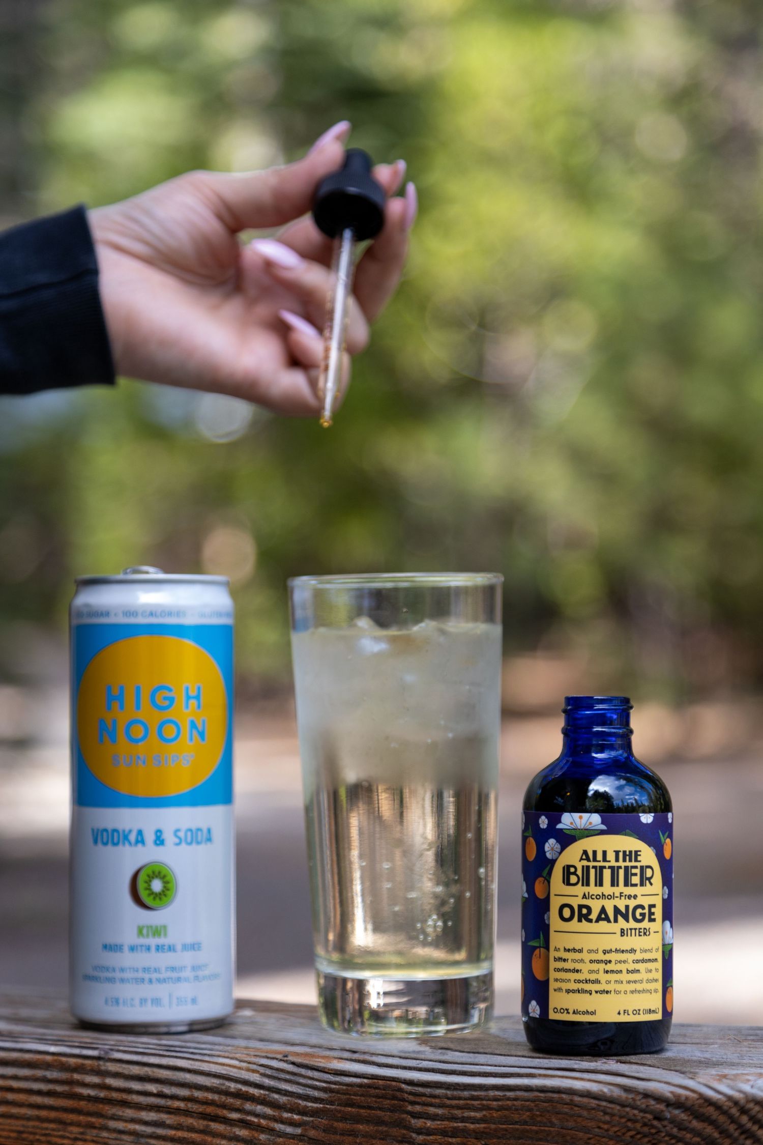 Orange bitters bottle and sparkling beverage on wooden railing with forested background in soft focus.