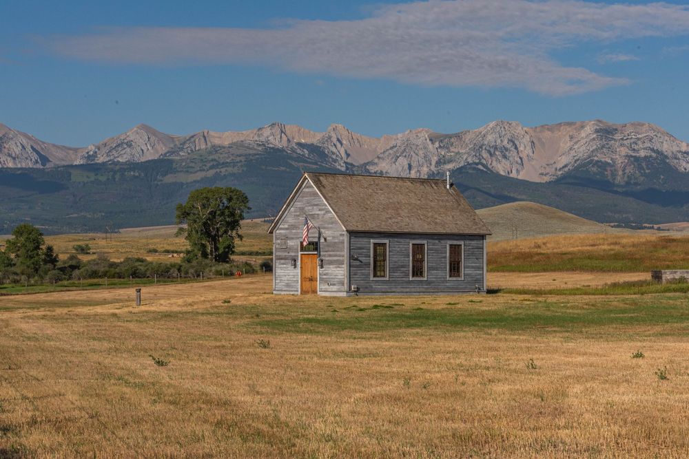 A rustic wooden house stands solitary in a golden prairie field with snow-capped mountains rising in the background.