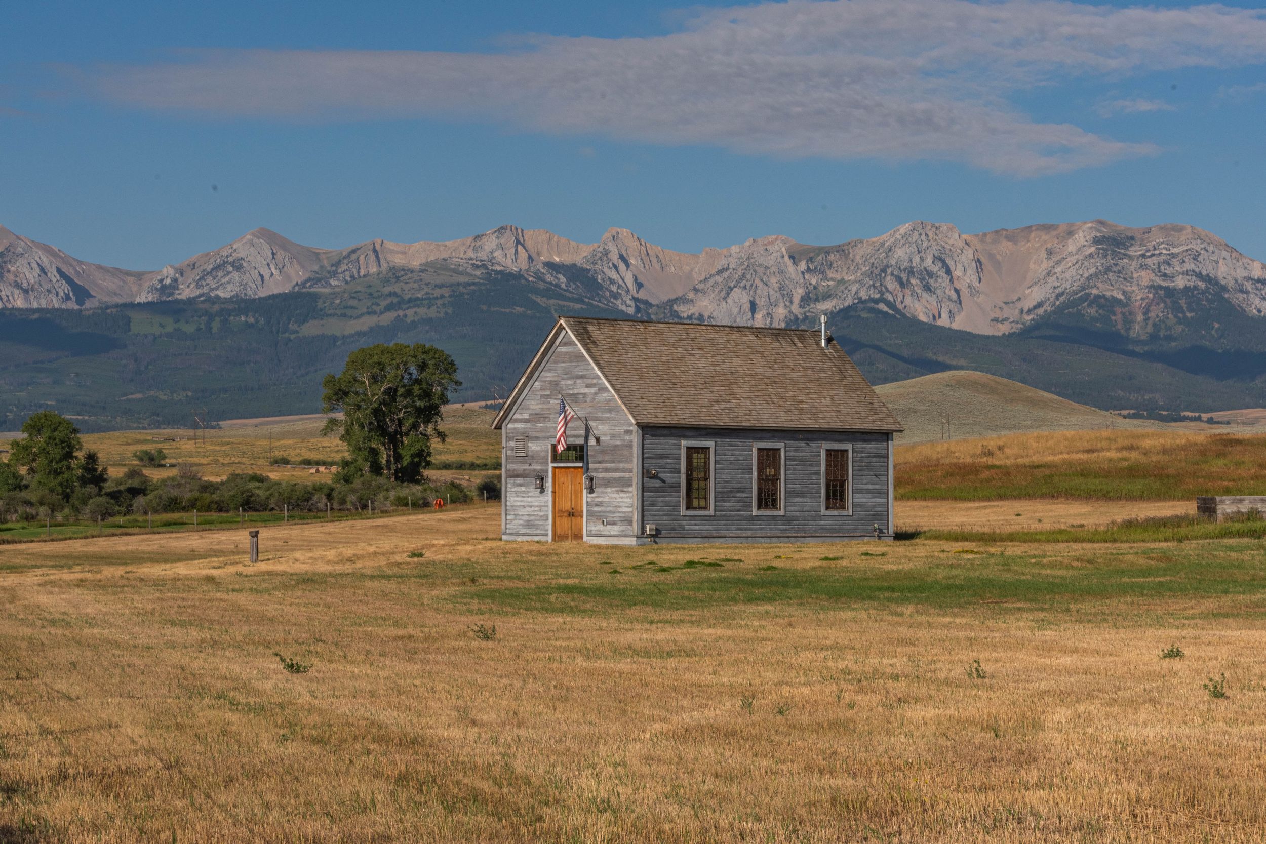 A rustic wooden house stands solitary in a golden prairie field with snow-capped mountains rising in the background.