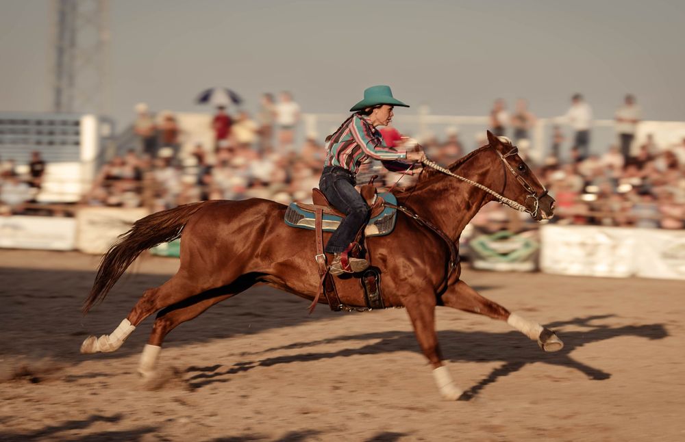 A cowgirl rides a chestnut horse at full gallop during a rodeo competition in a sandy arena.