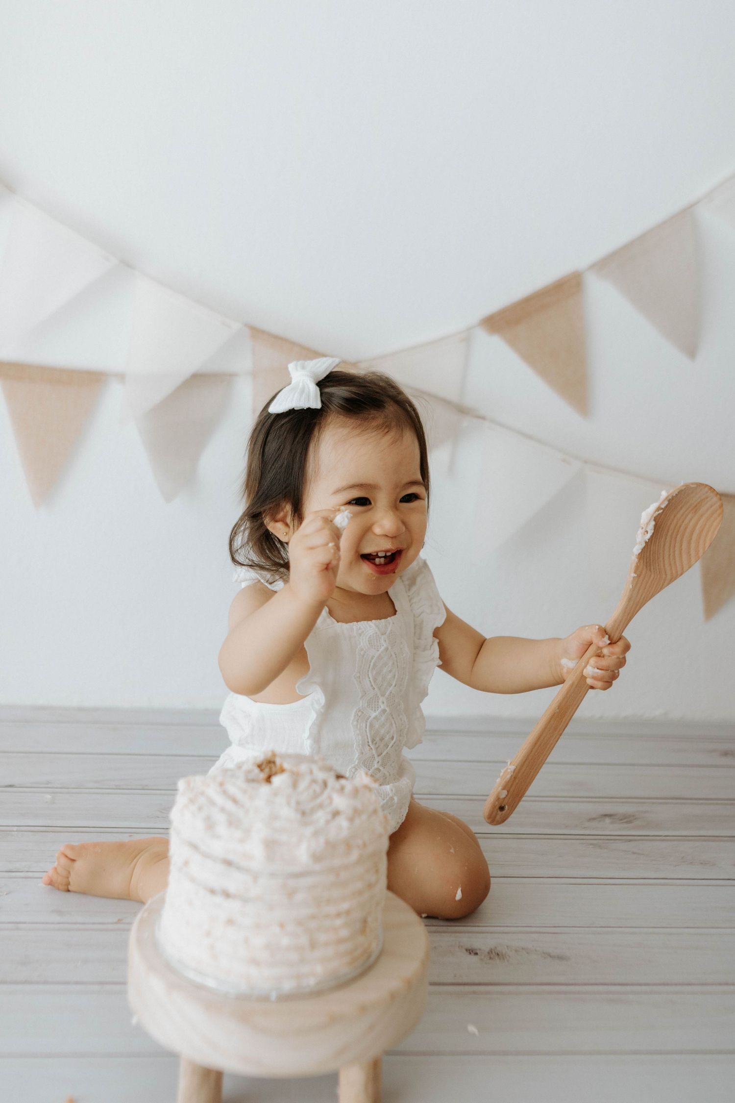 A series of four photos showing a toddler in a white lacy dress sitting on a white floor with bunting decorations above. Cake in front