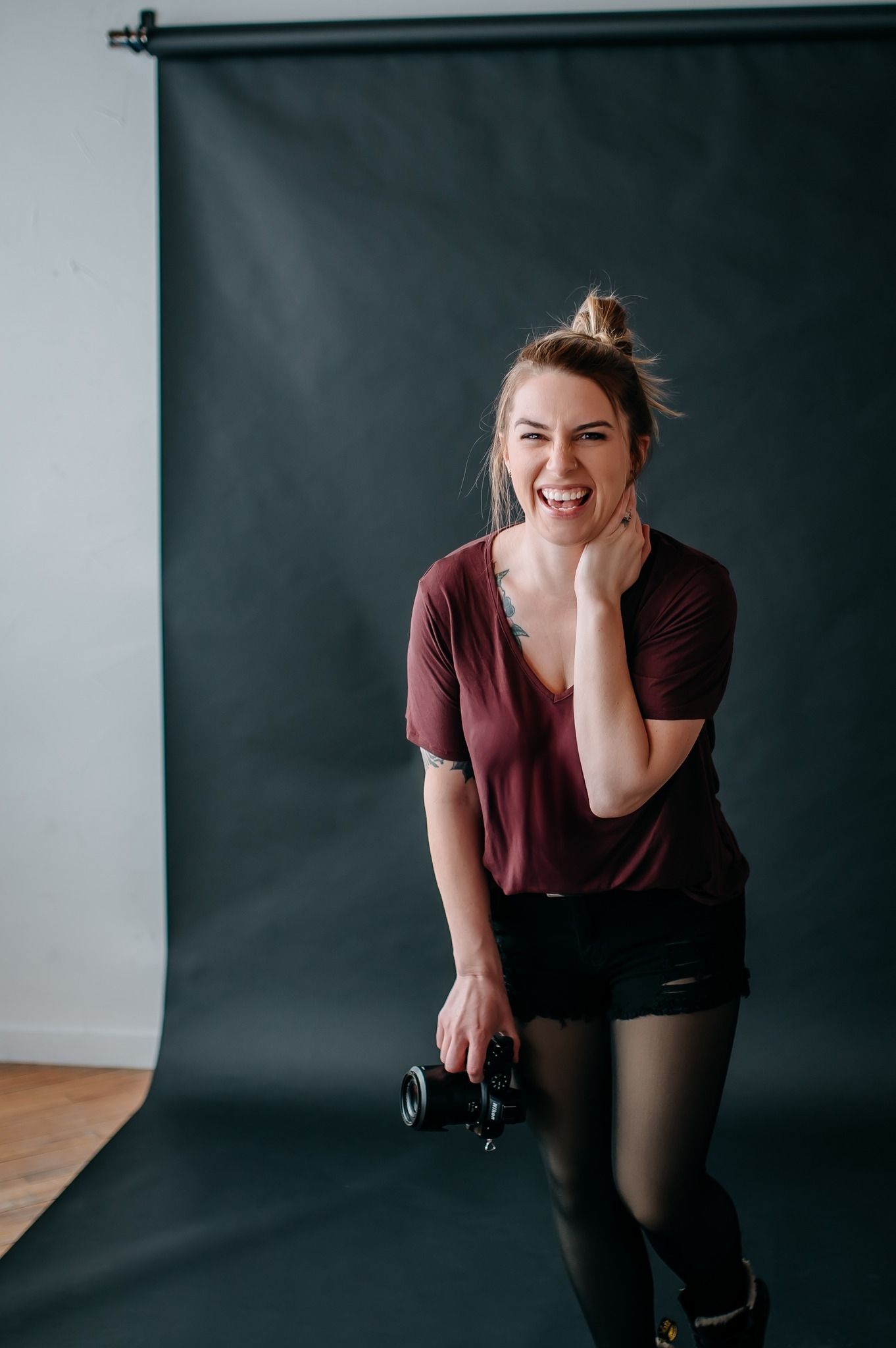 A photographer in burgundy shirt and black shorts laughs joyfully while holding her nikon camera against a dark backdrop.