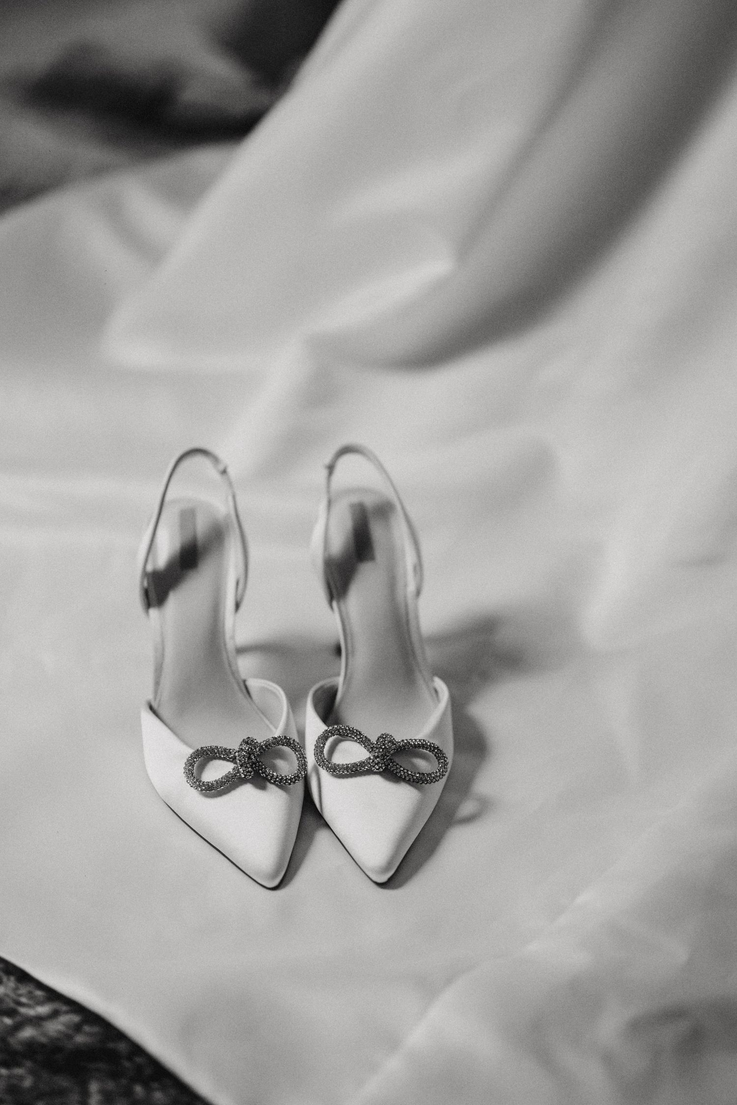 A pair of elegant white bridal shoes with bow details sits atop a soft fabric in black and white photography.
