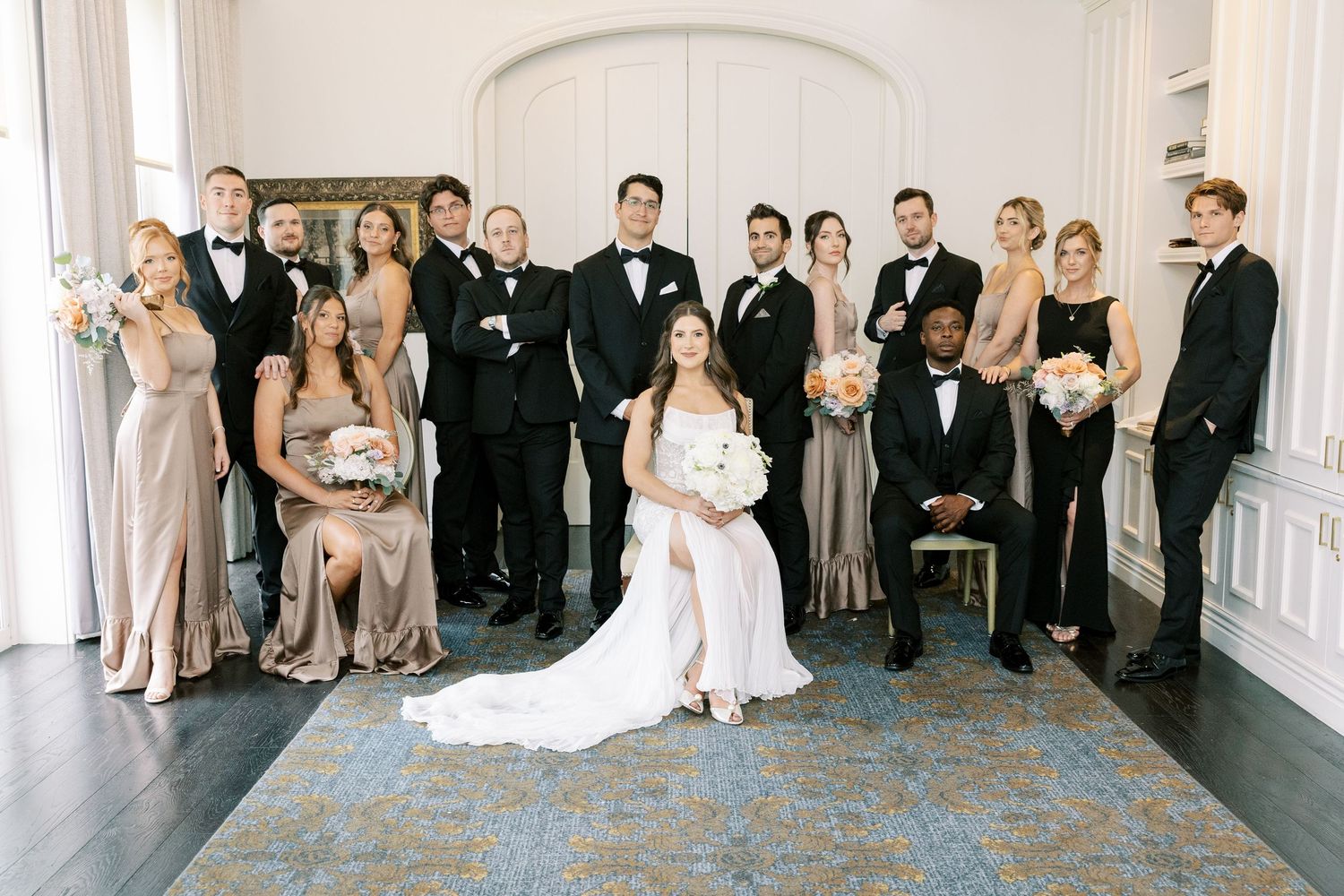 Wedding party wearing formal attire poses together on blue patterned carpet in elegant white room.