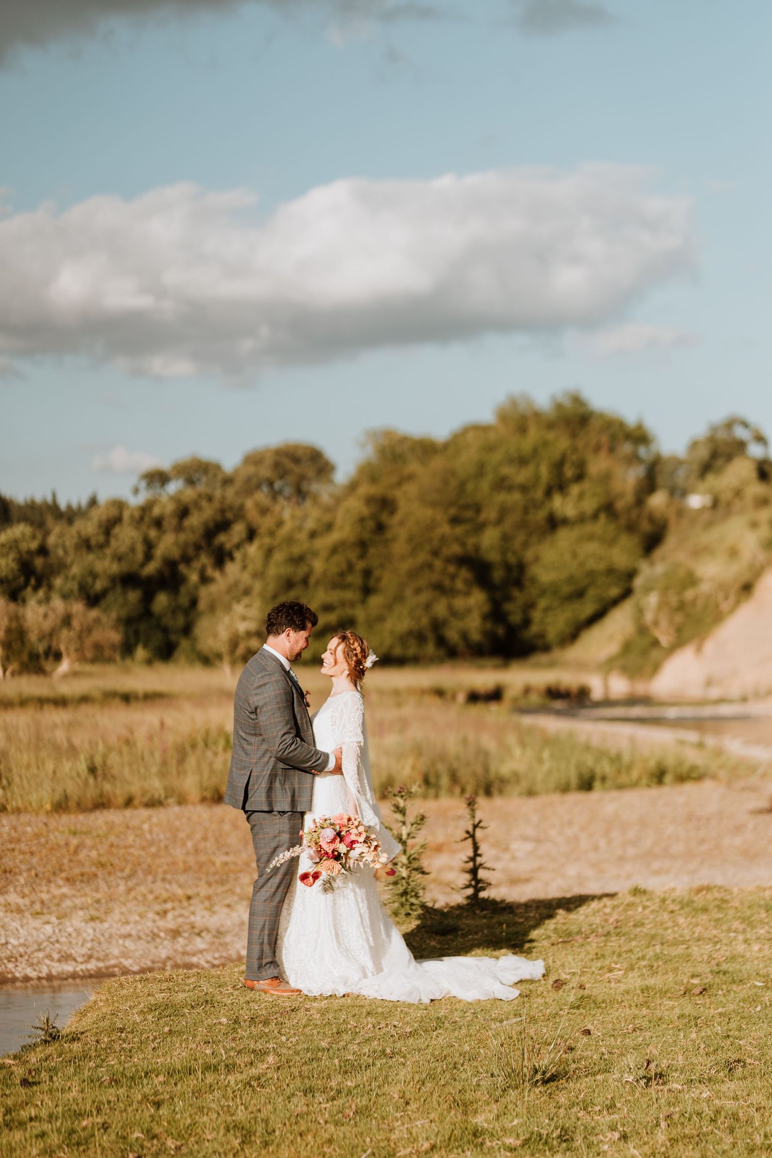 Couple in wedding attire standing together on a grassy hill with autumn trees and cloudy sky in the background.