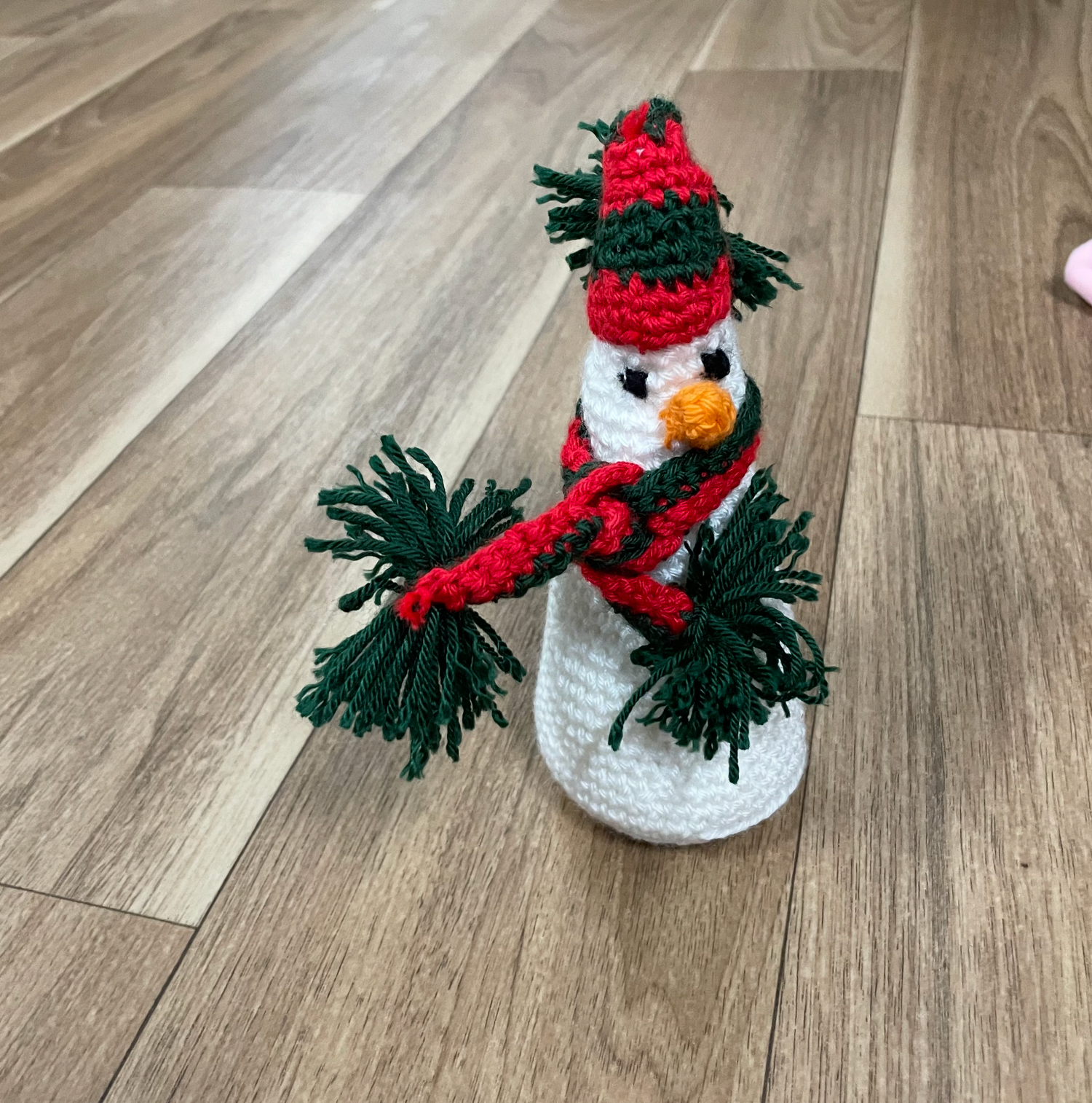 A crocheted snowman with green pom poms and red winter accessories on hardwood floor.