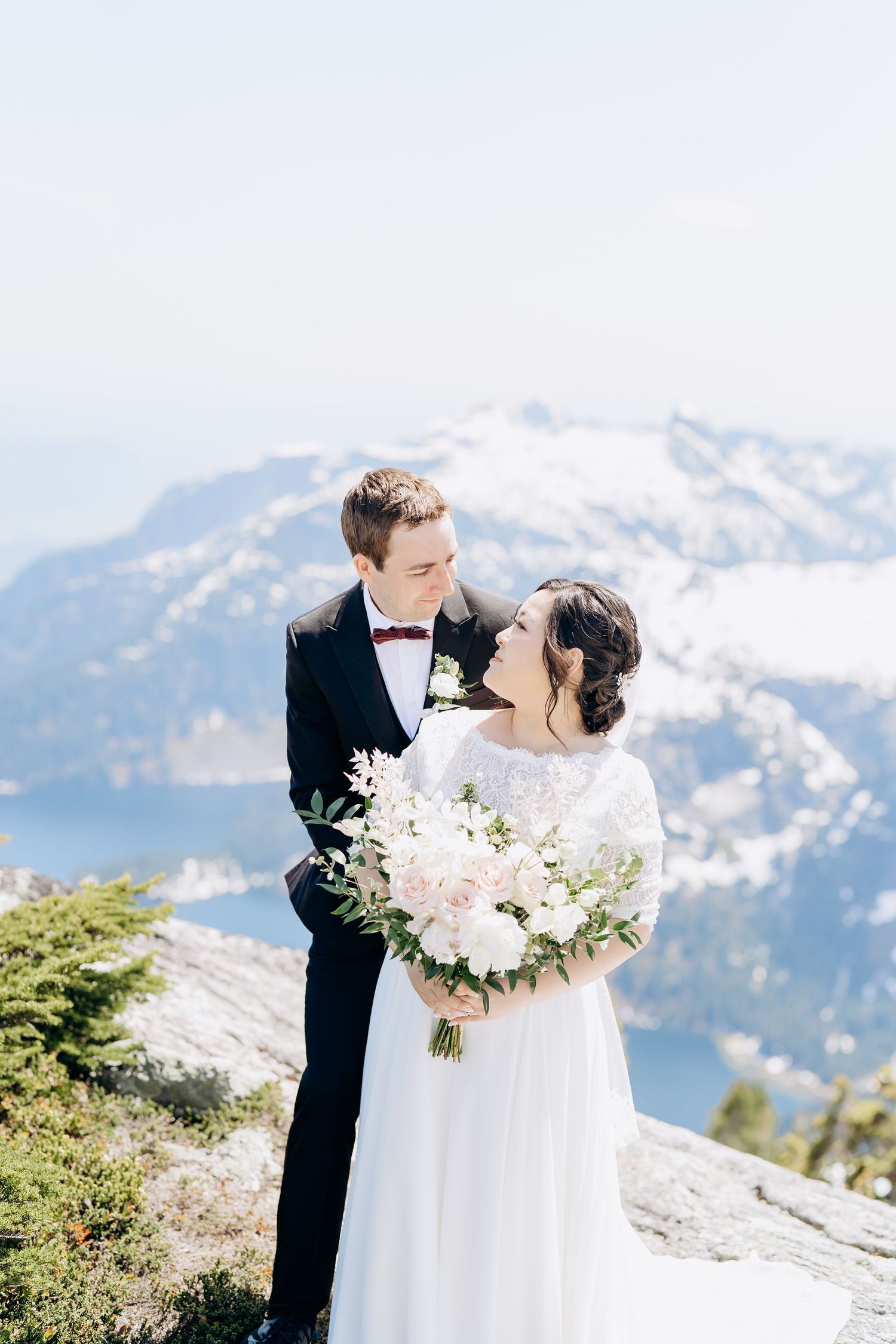 A romantic wedding couple embraces on a snowy mountaintop with stunning snow-capped peaks in the background.