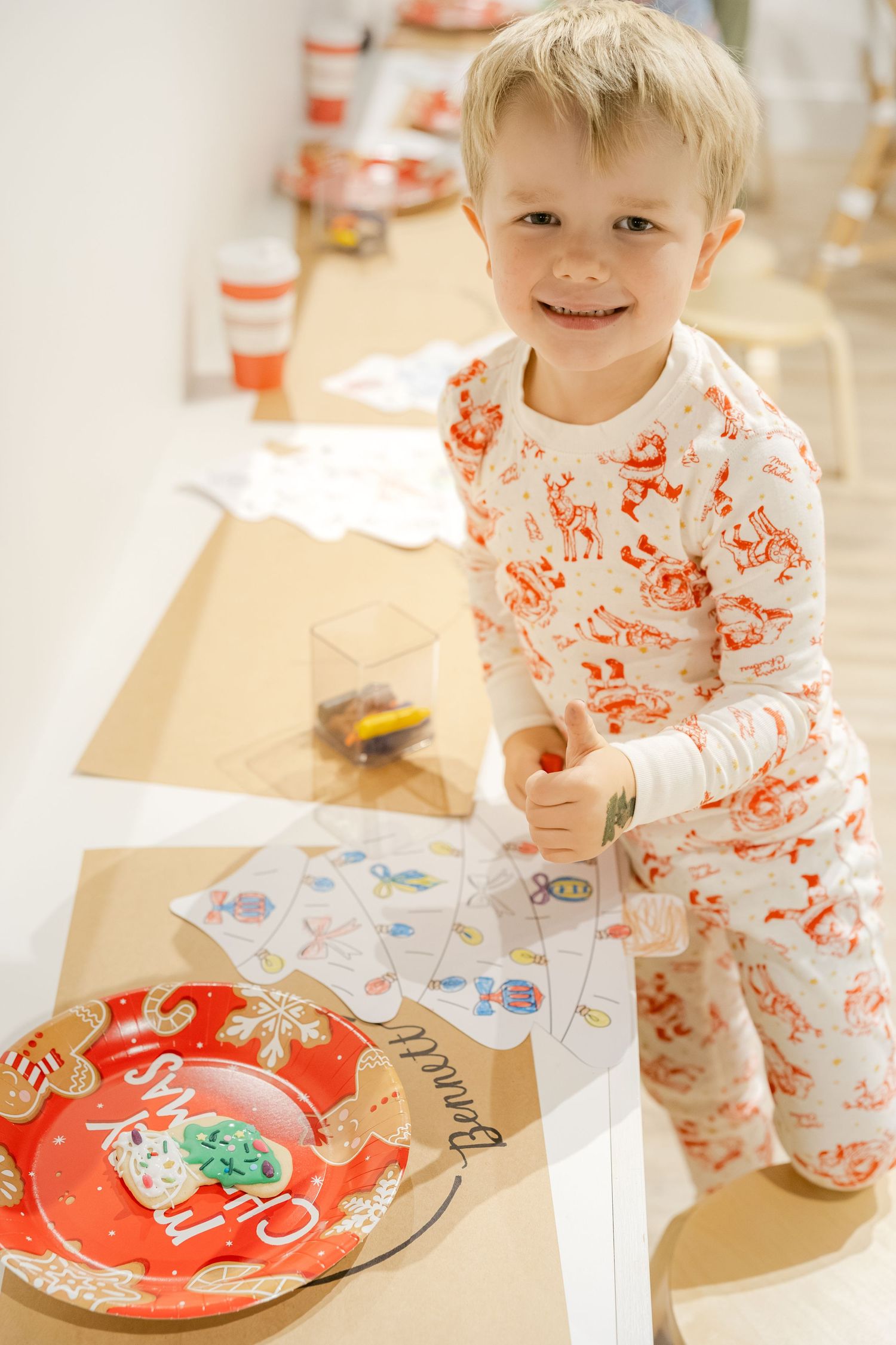 A small child in christmas pajamas decorates holiday cookies and colors christmas artwork at a table.