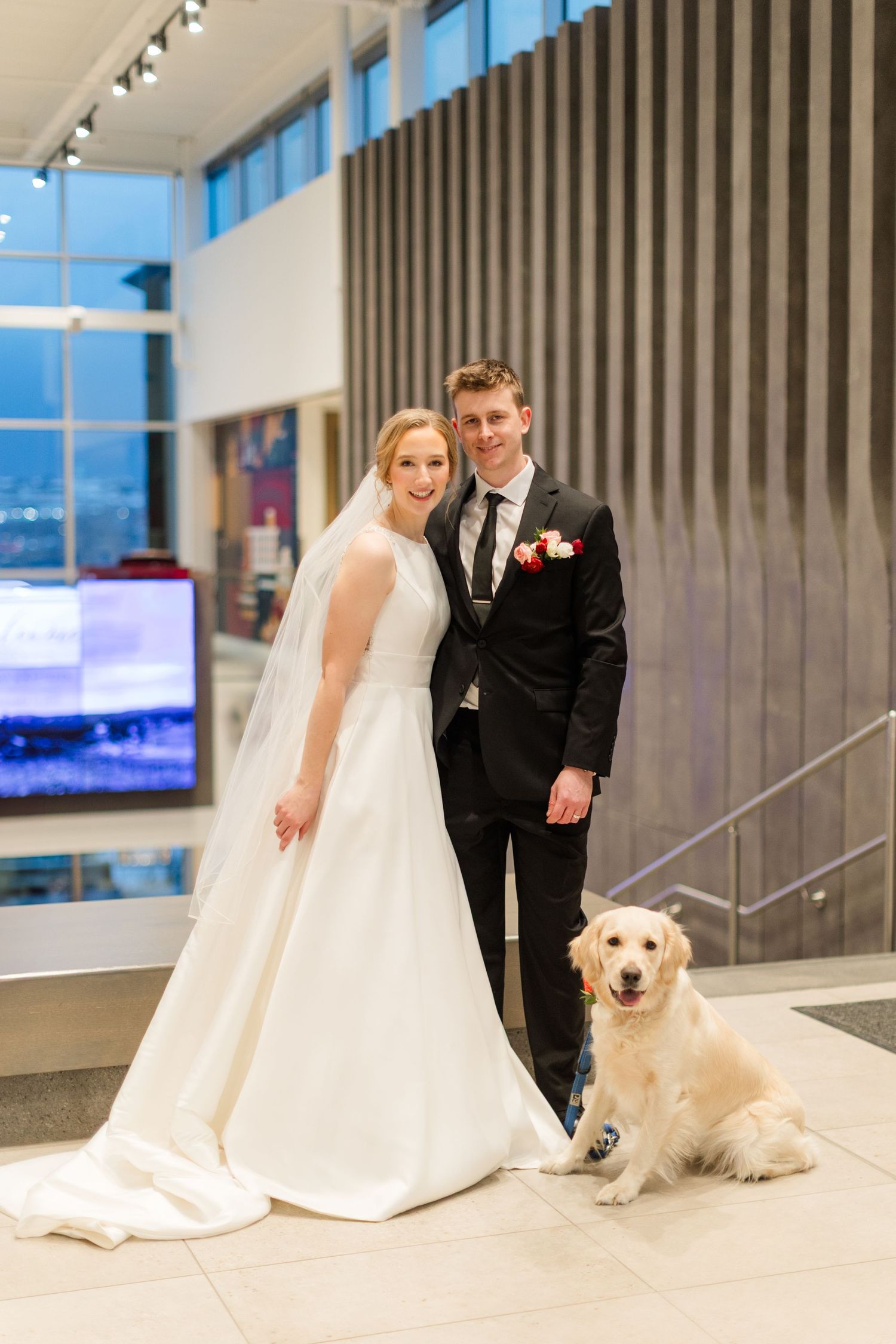 A bride and groom pose with their golden retriever in a modern indoor venue with glass windows and vertical panels.
