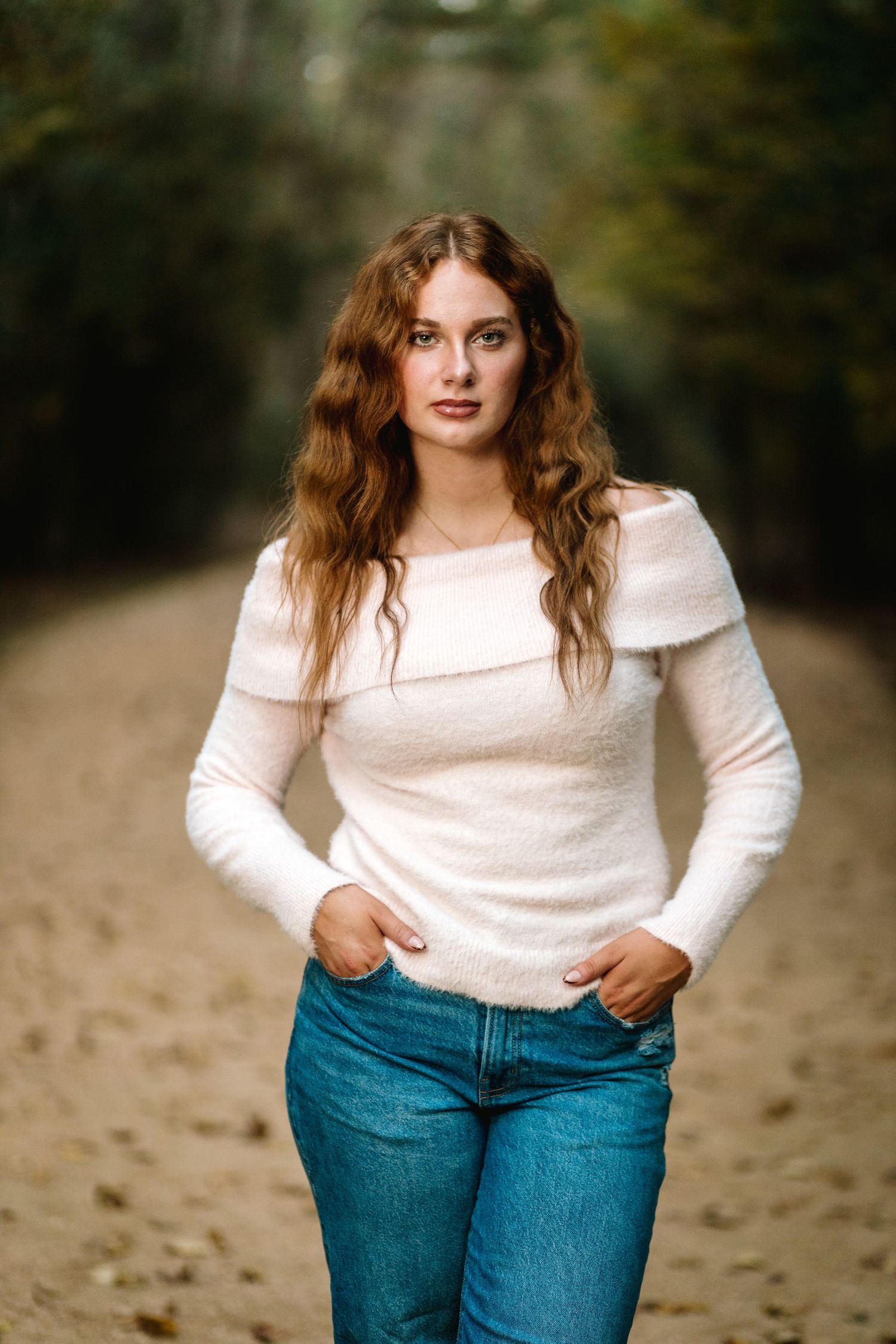 A person wearing a white sweater and blue jeans poses casually outdoors in a natural setting.
