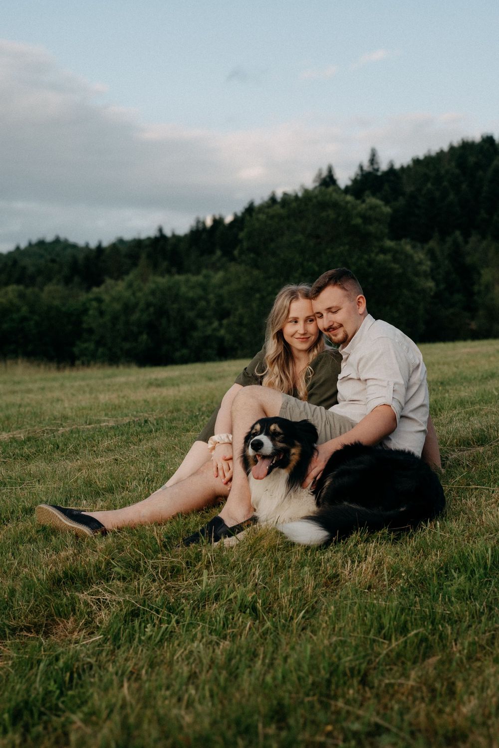 A couple relaxes with their dog in a sunny meadow surrounded by forest.