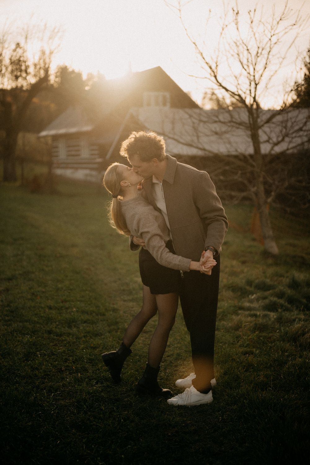 A couple shares a tender moment at sunset in a grassy field with a house in the background.