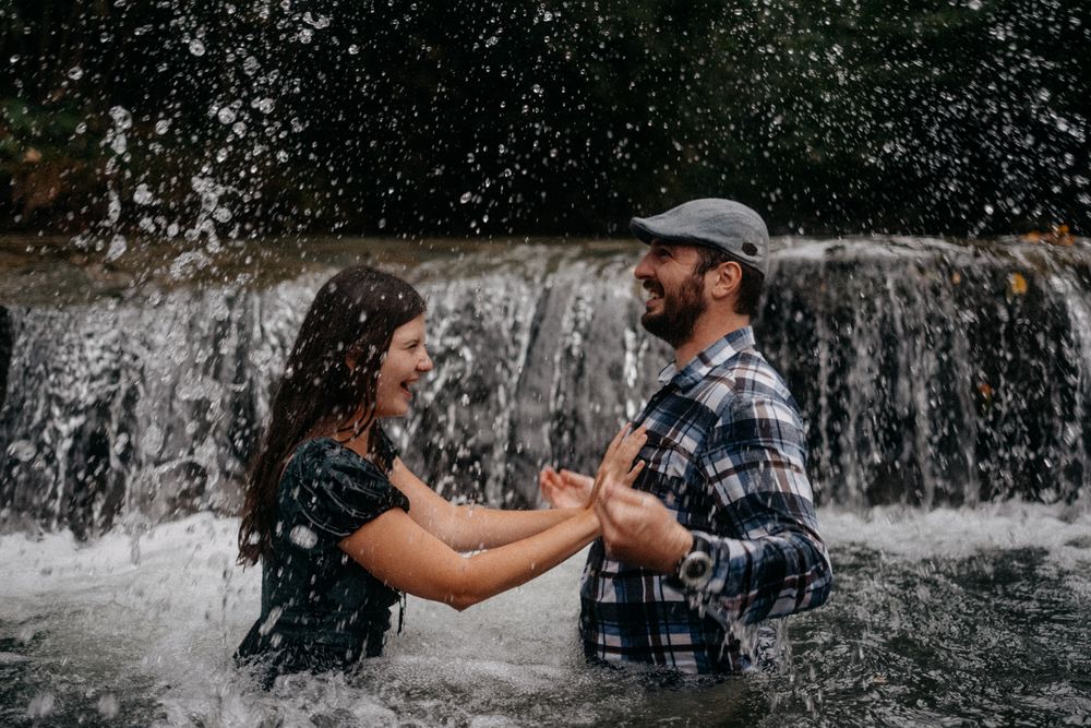 Two people splash and play in a waterfall while wearing casual clothes and having fun on a summer day.