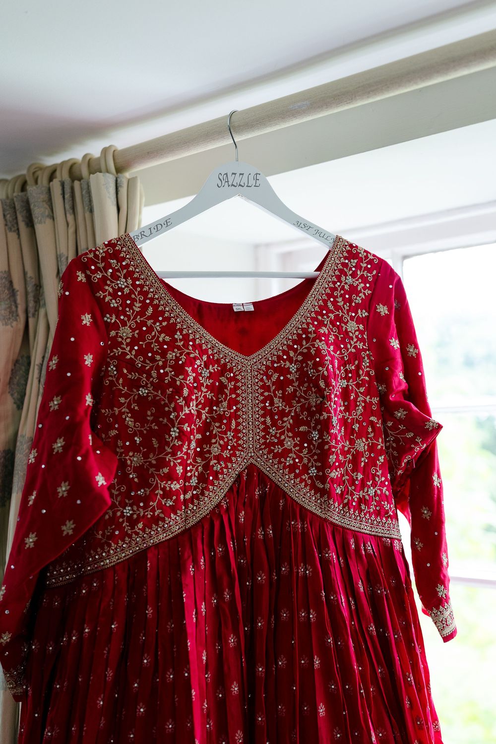 Red velvet dress with intricate gold embroidery and beading displayed on a hanger against white wall.