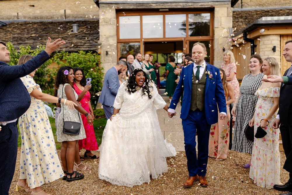 Wedding party gathers outside a rustic stone building for formal group photos on a sunny afternoon.
