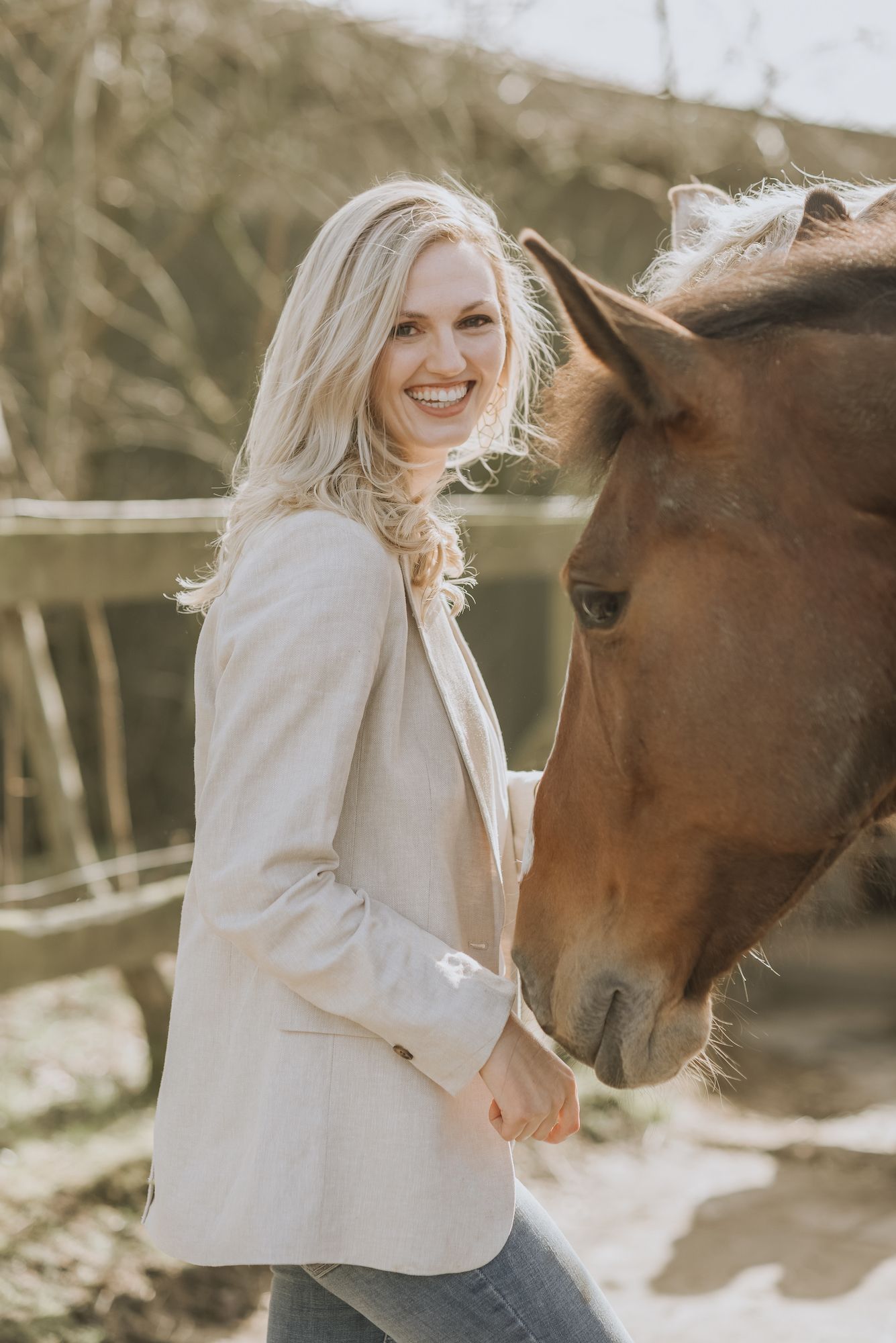Woman in beige blazer standing next to brown horse in natural sunlit setting.