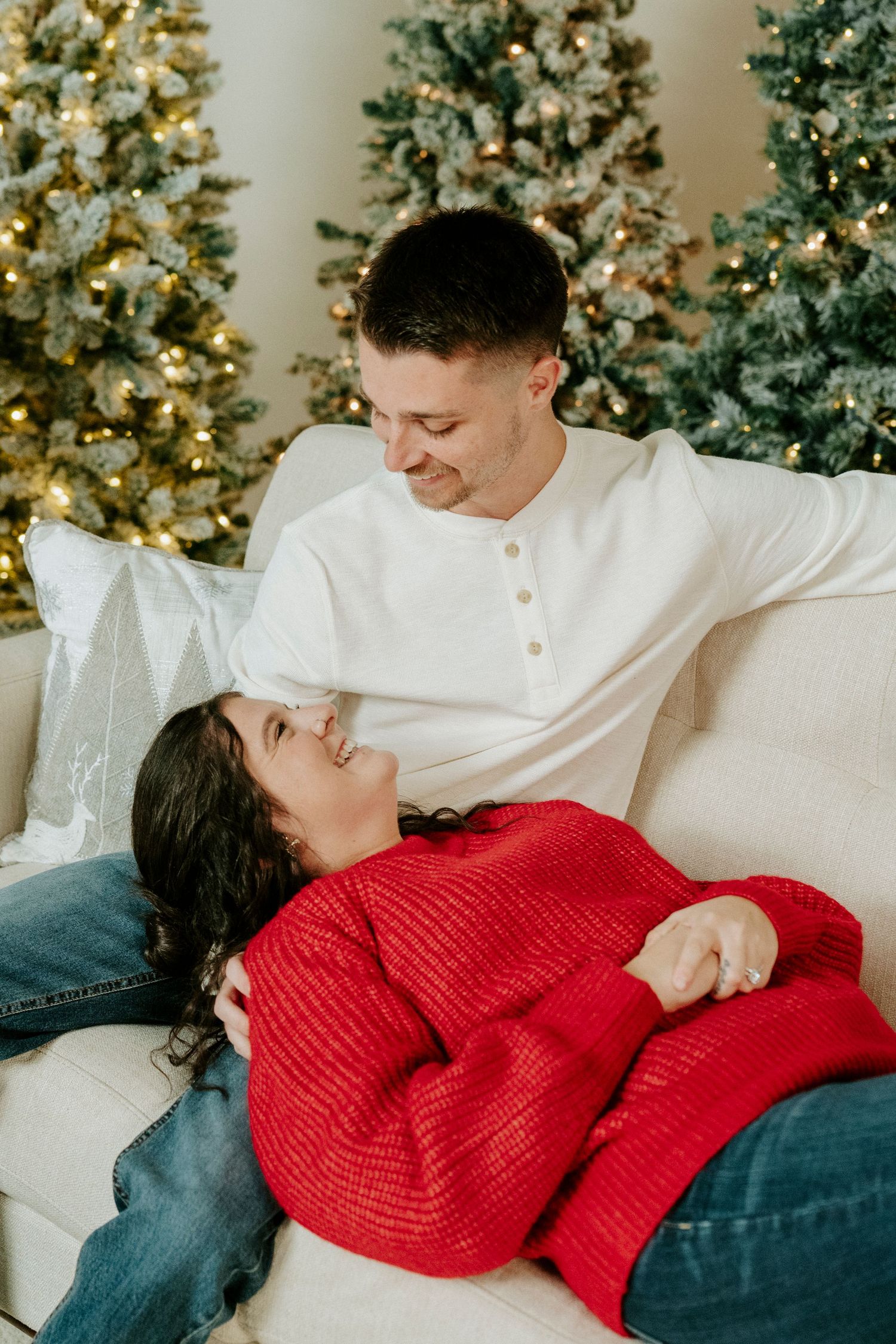 A couple in white and red sweaters share an intimate moment on a couch with Christmas trees in the background.