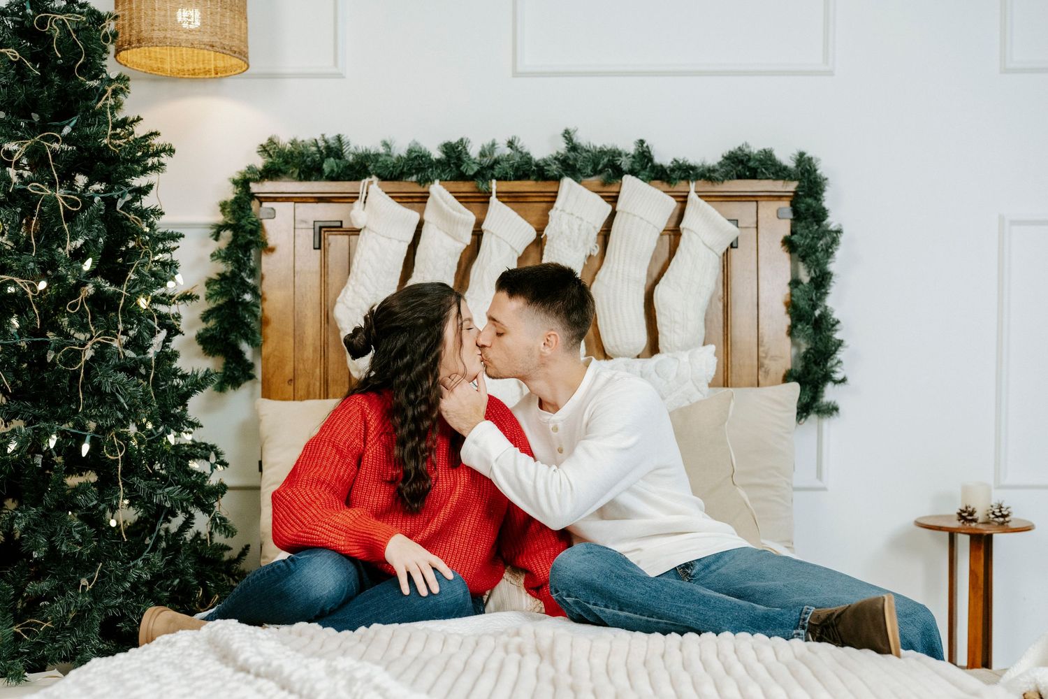 Couple in red and white sweaters share romantic moment on bed with Christmas stockings and tree in background.