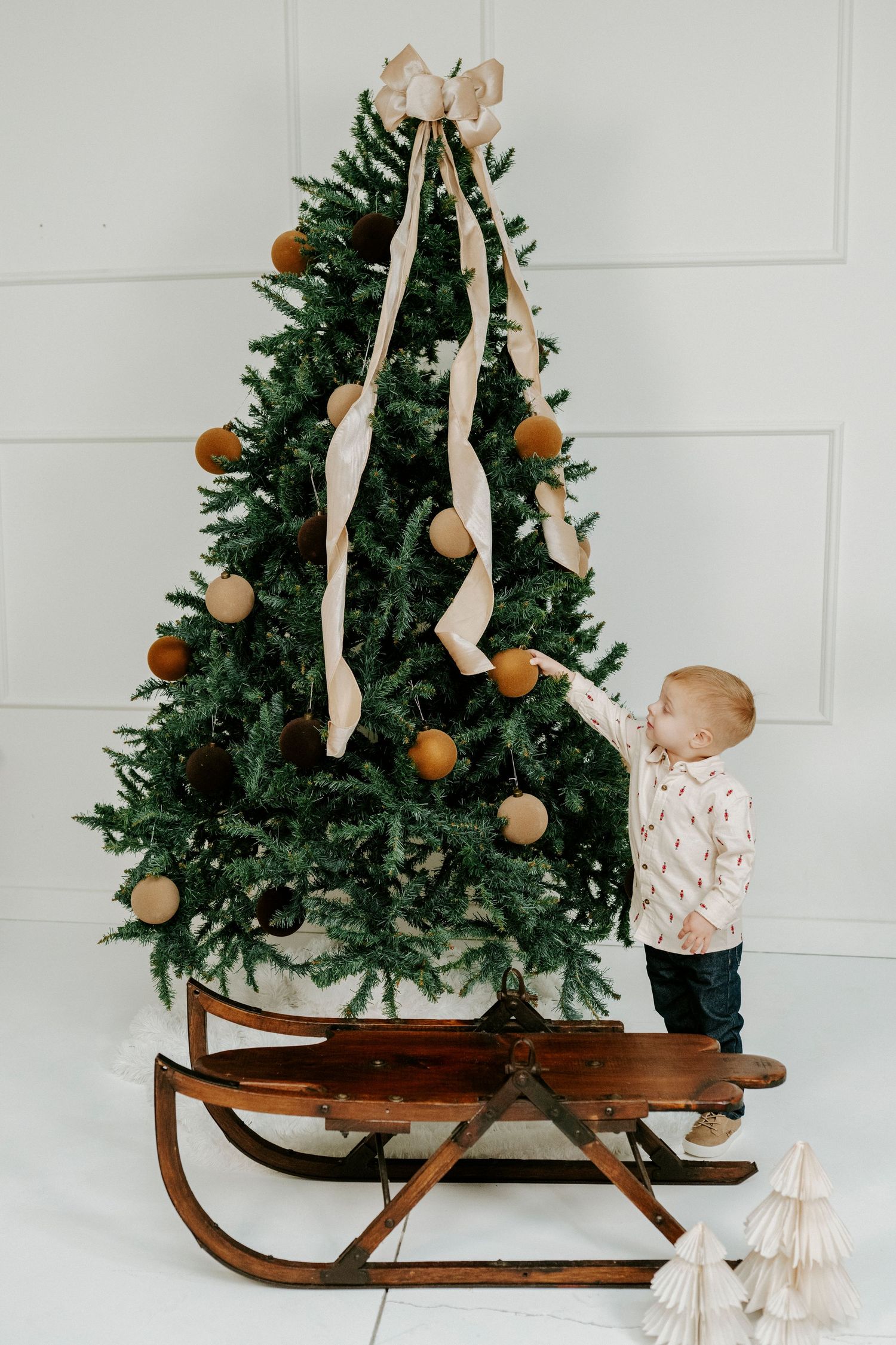 Small child decorates a Christmas tree placed on vintage wooden sleigh against white wall.