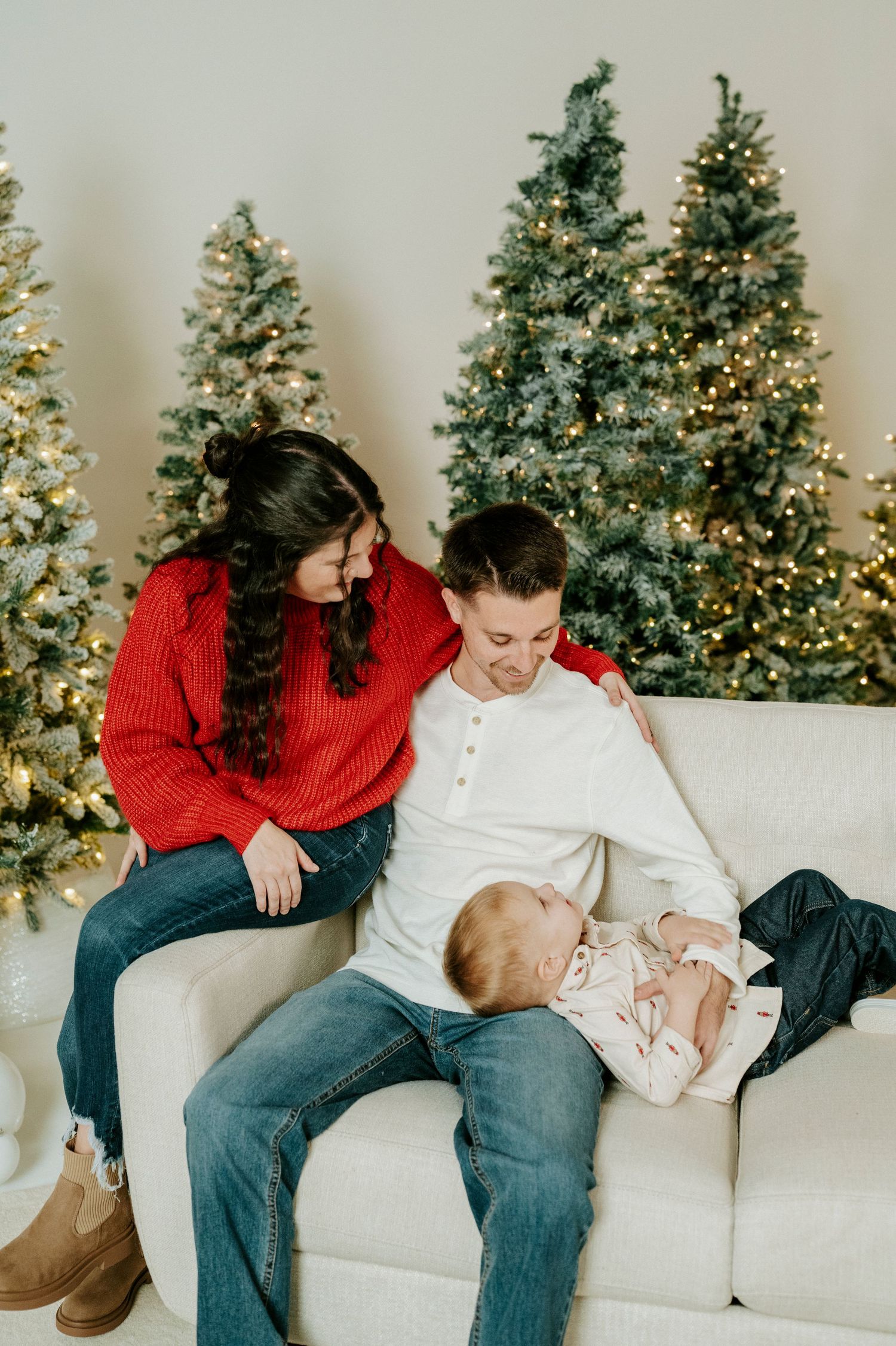 A couple in red and white sweaters share playful moments on a white couch with decorated Christmas trees in background.