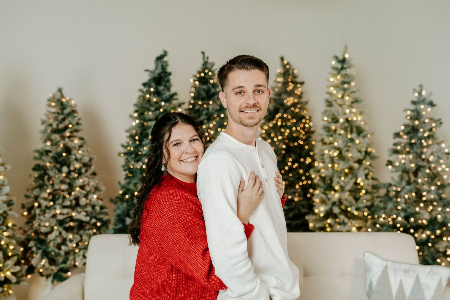 A couple poses together in front of illuminated Christmas trees wearing red and white sweaters.