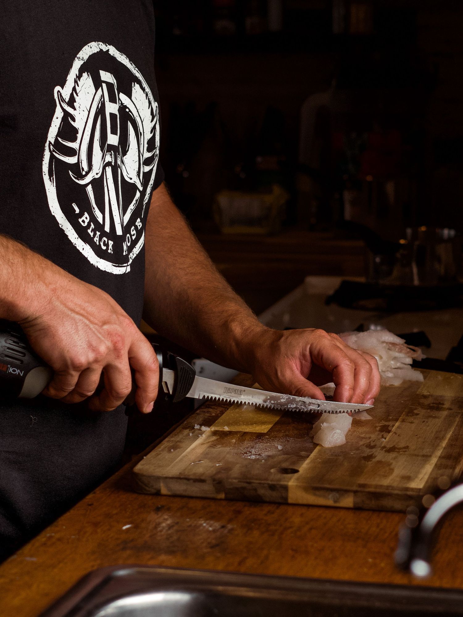 Hands using a knife on a wooden cutting board with a logo visible on dark clothing.