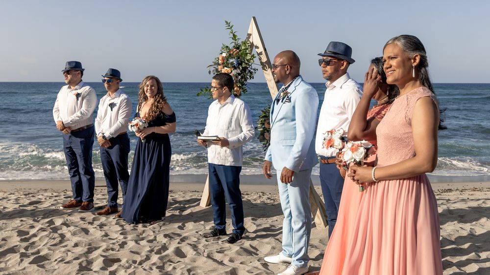 A group gathers on a sunny beach for a wedding ceremony with the ocean waves in the background.
