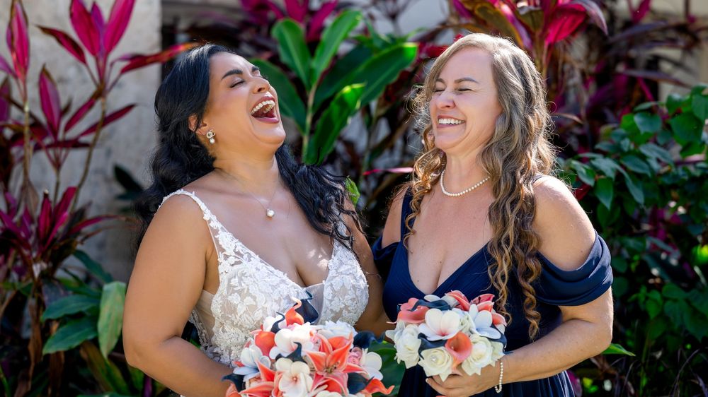 Two women in wedding attire laughing joyfully together in a garden setting with colorful tropical plants.