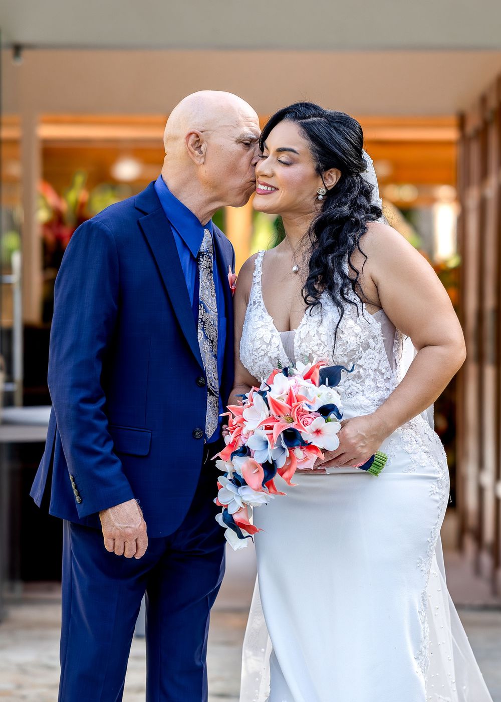 A couple in wedding attire sharing romantic moments in an outdoor corridor with pink floral bouquet.