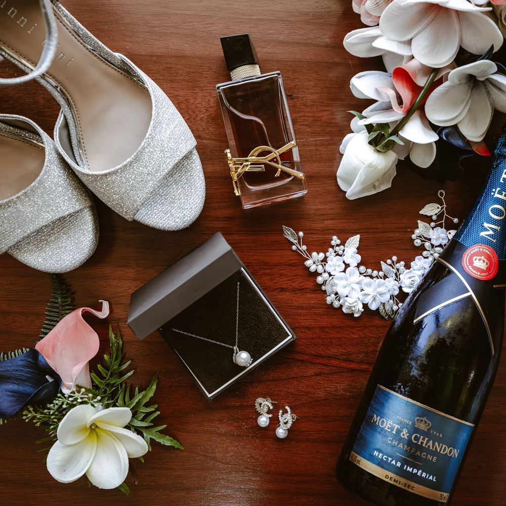 Wedding accessories including white shoes, flowers, and champagne arranged on wooden surface.