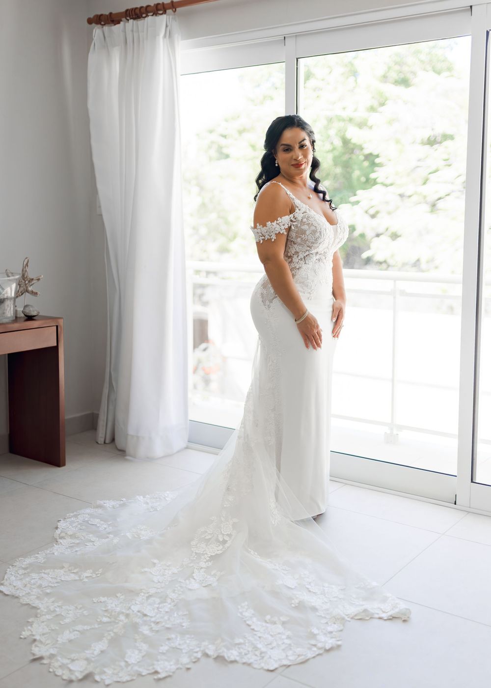 A beautiful wedding dress train with intricate lace details spreads across a bright white floor in a sunlit room.