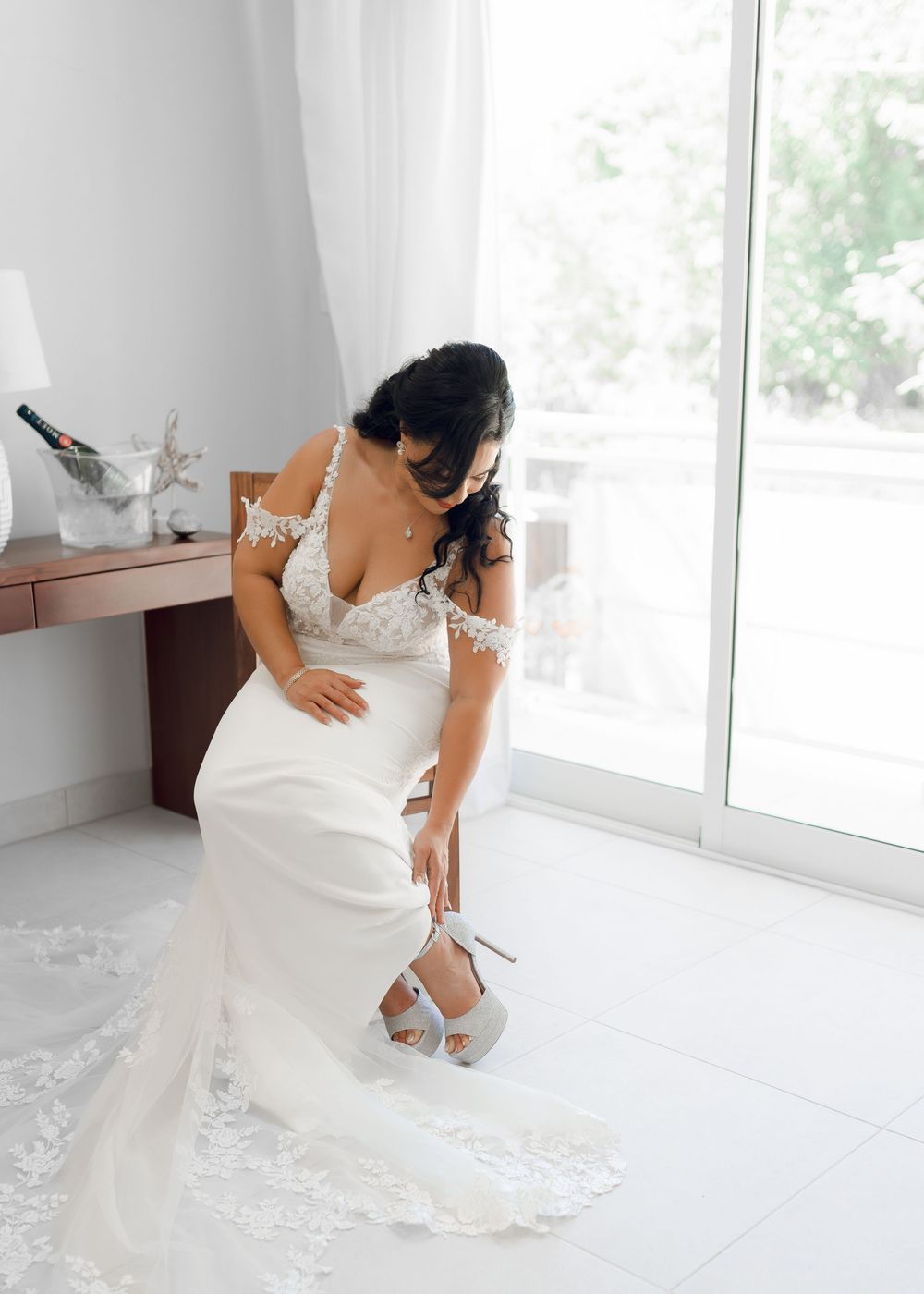 Bride in white wedding gown adjusting her dress in a bright sunlit room with white decor.