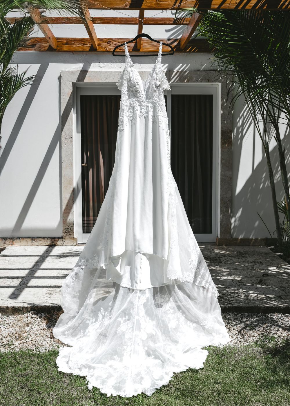 A line of white wedding dresses displayed against a white wall with palm tree shadows.