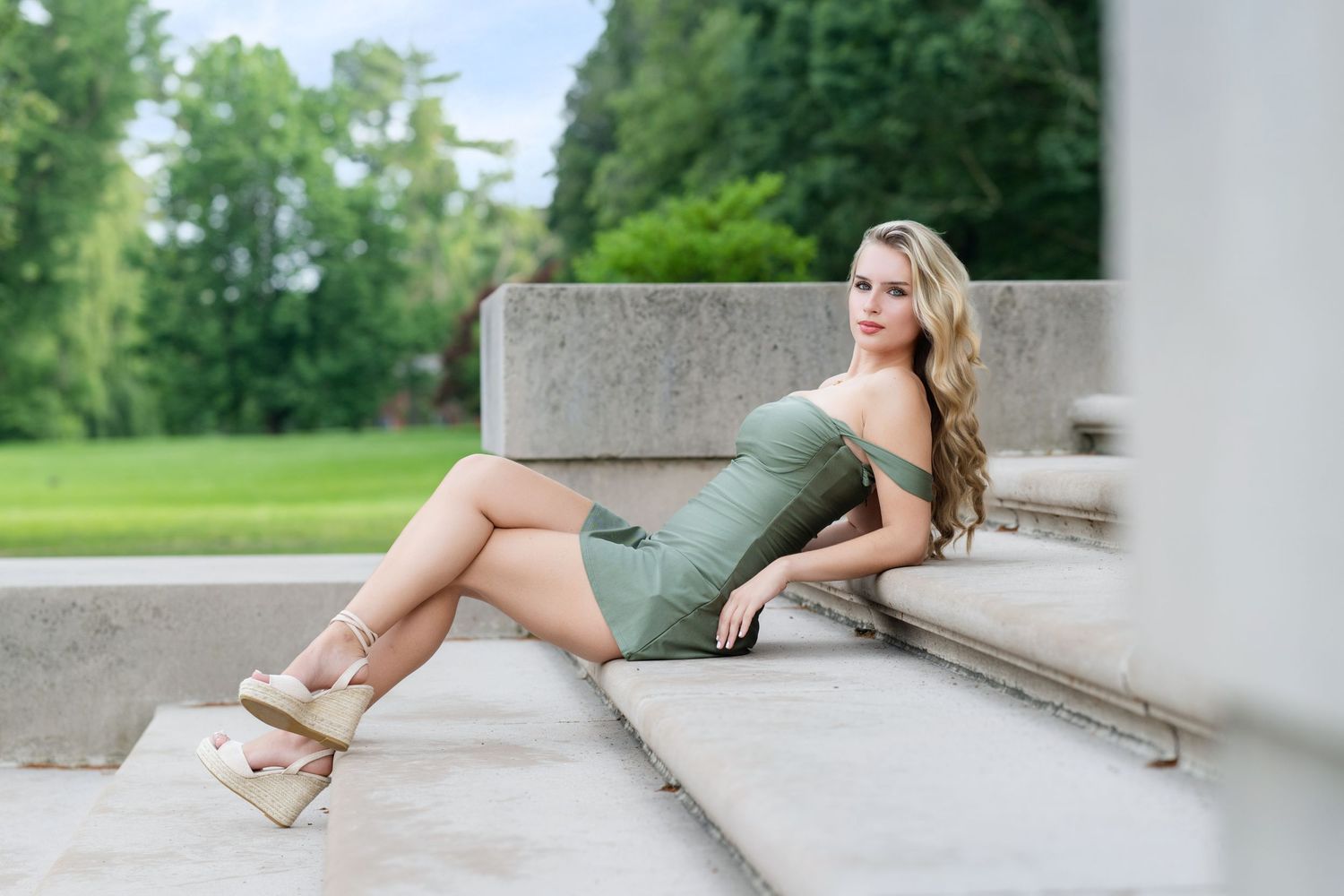 Woman in olive green dress poses elegantly on concrete steps outdoors.
