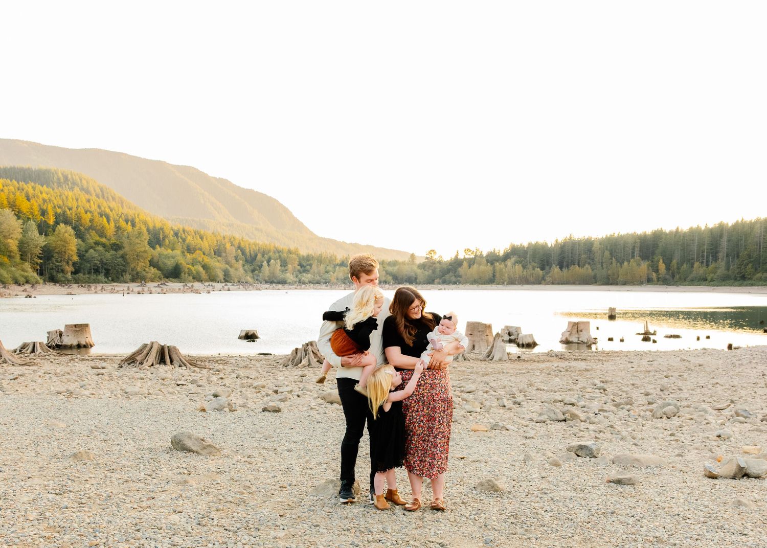 A family shares a tender moment together on a rocky beach shore with mountains and forest in the background.