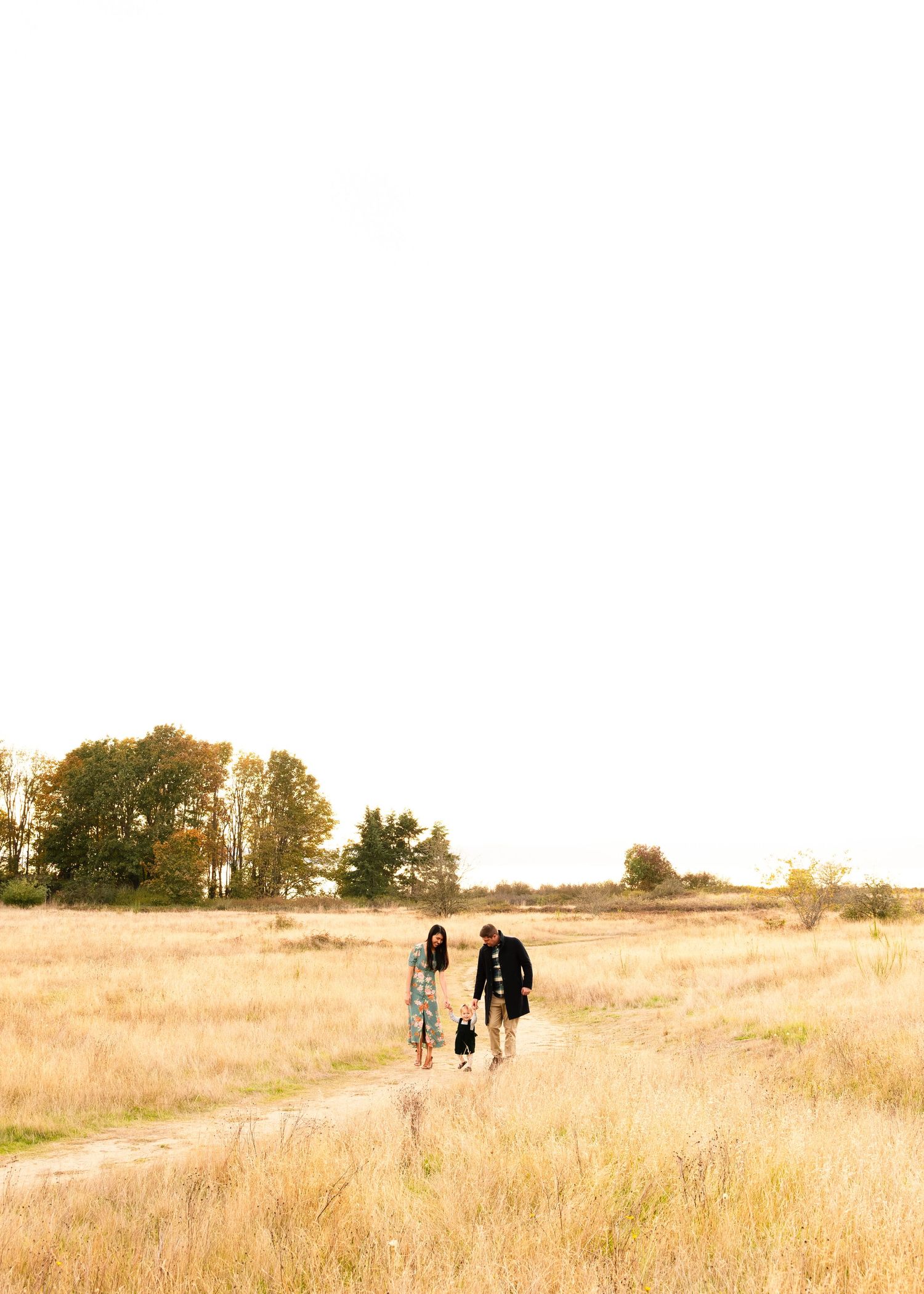 People walking on a winding dirt path through golden grass fields at sunset with trees in the distance.