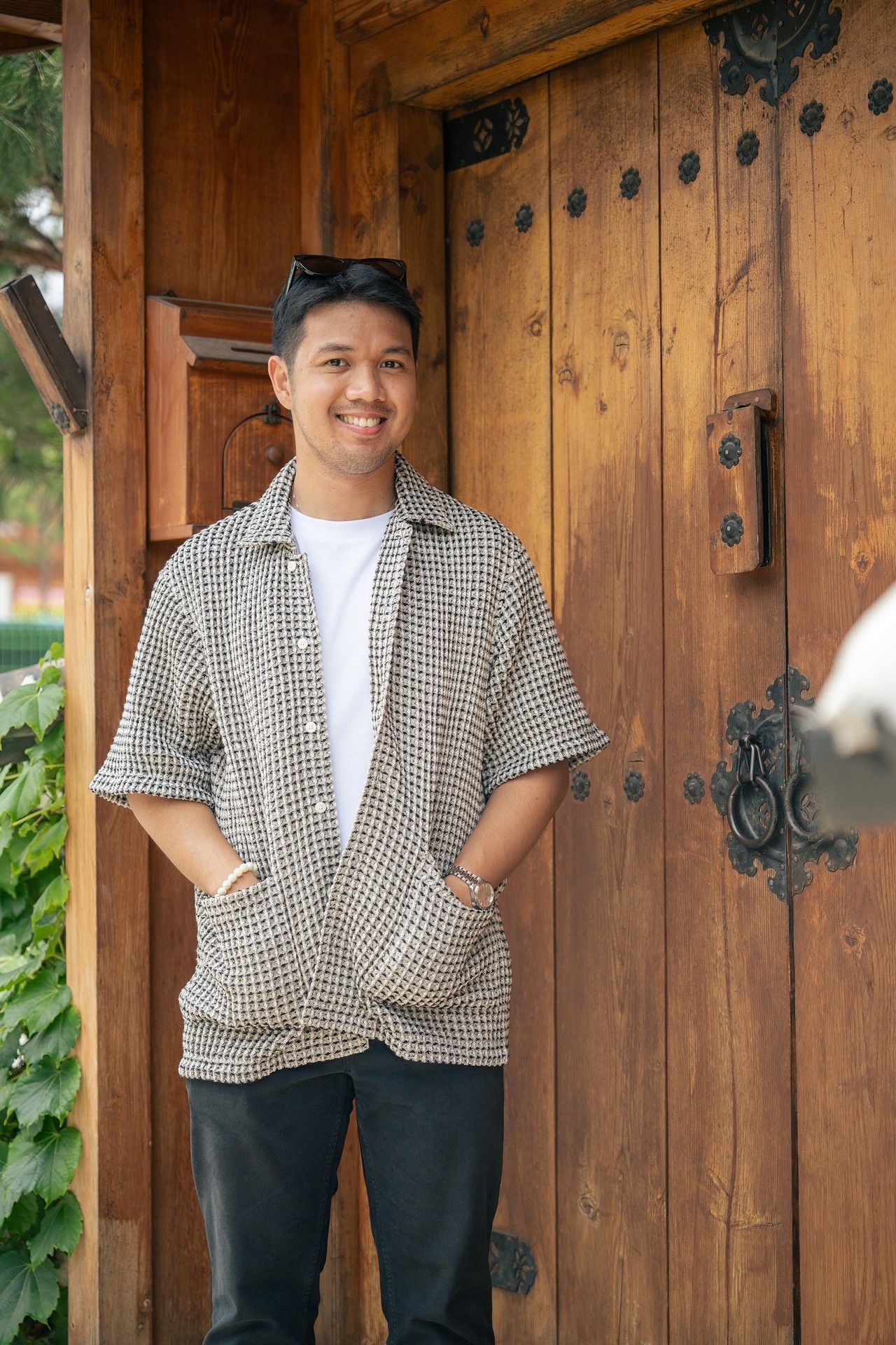 A person in a checkered shirt and white t-shirt poses against a rustic wooden door in a casual outdoor setting.