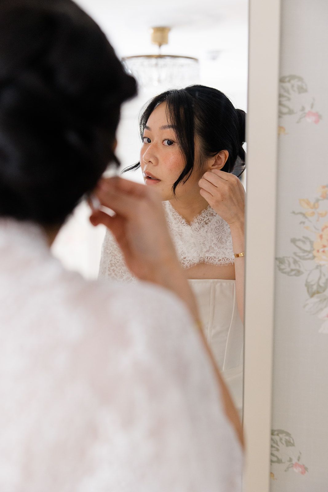 A bride in a white lace dress looks into a mirror while getting ready for her wedding ceremony.