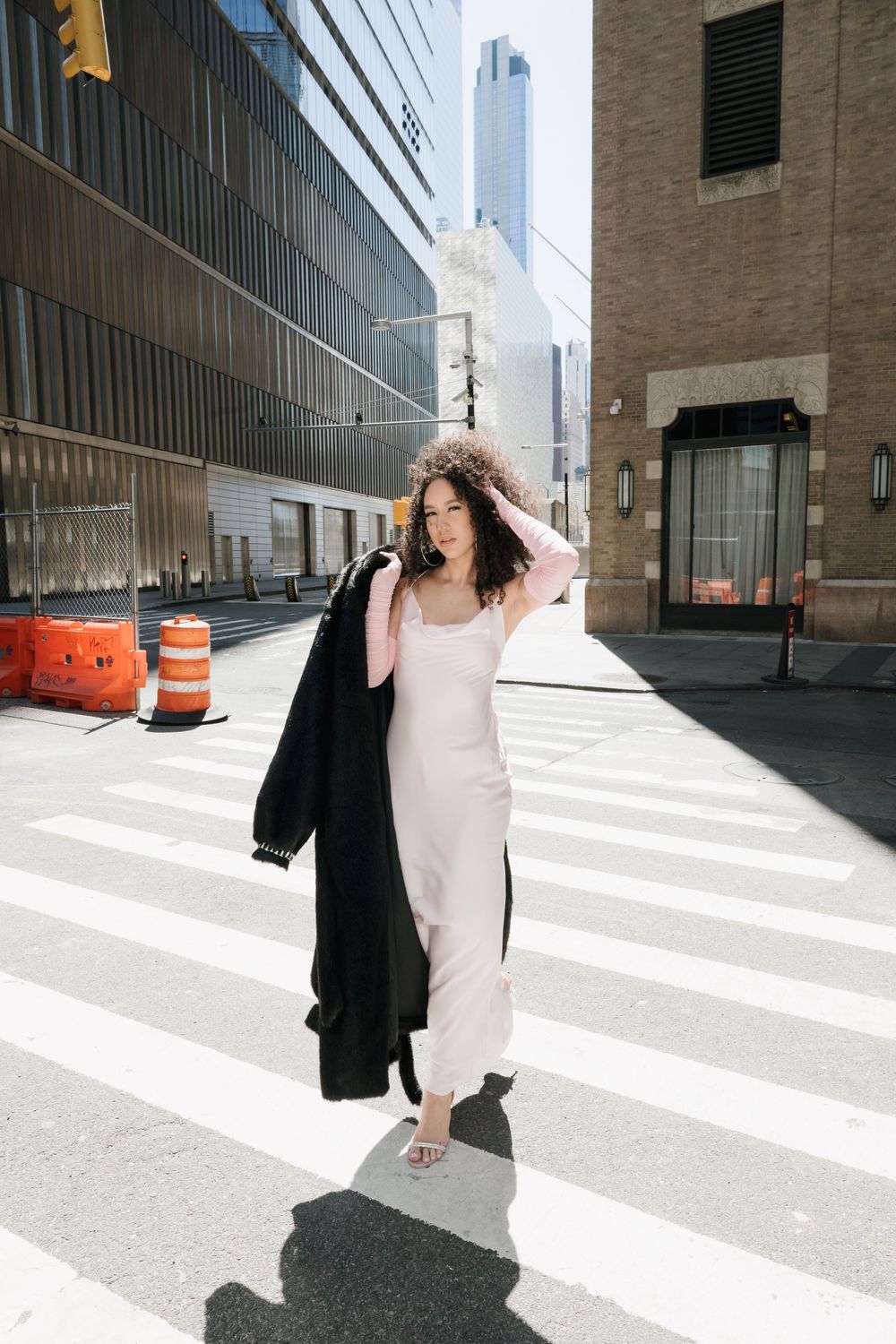 Woman in white slip dress carrying black coat walks across urban crosswalk on sunny day.