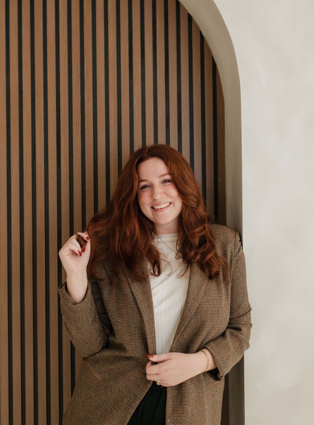 Person in brown blazer and white top posing against wooden slat wall.